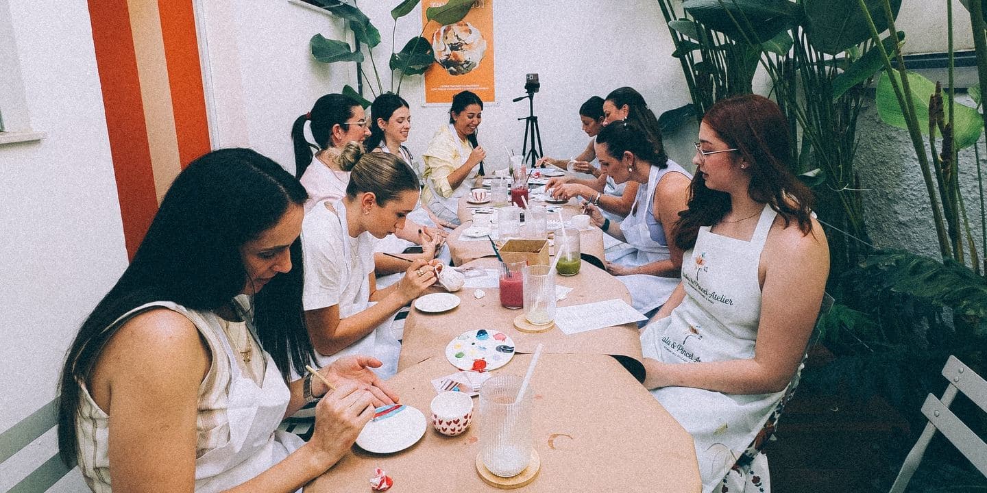 A mesa comprida do ateliê no quintal dos fundos, cheia de convidadas, embaixo das plantas