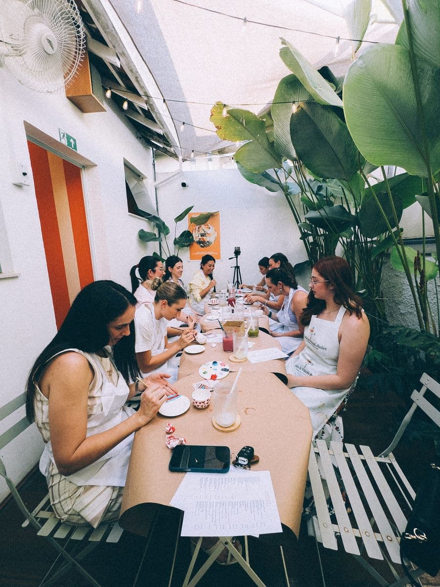 A mesa comprida do ateliê no quintal dos fundos, cheia de convidadas, embaixo das plantas